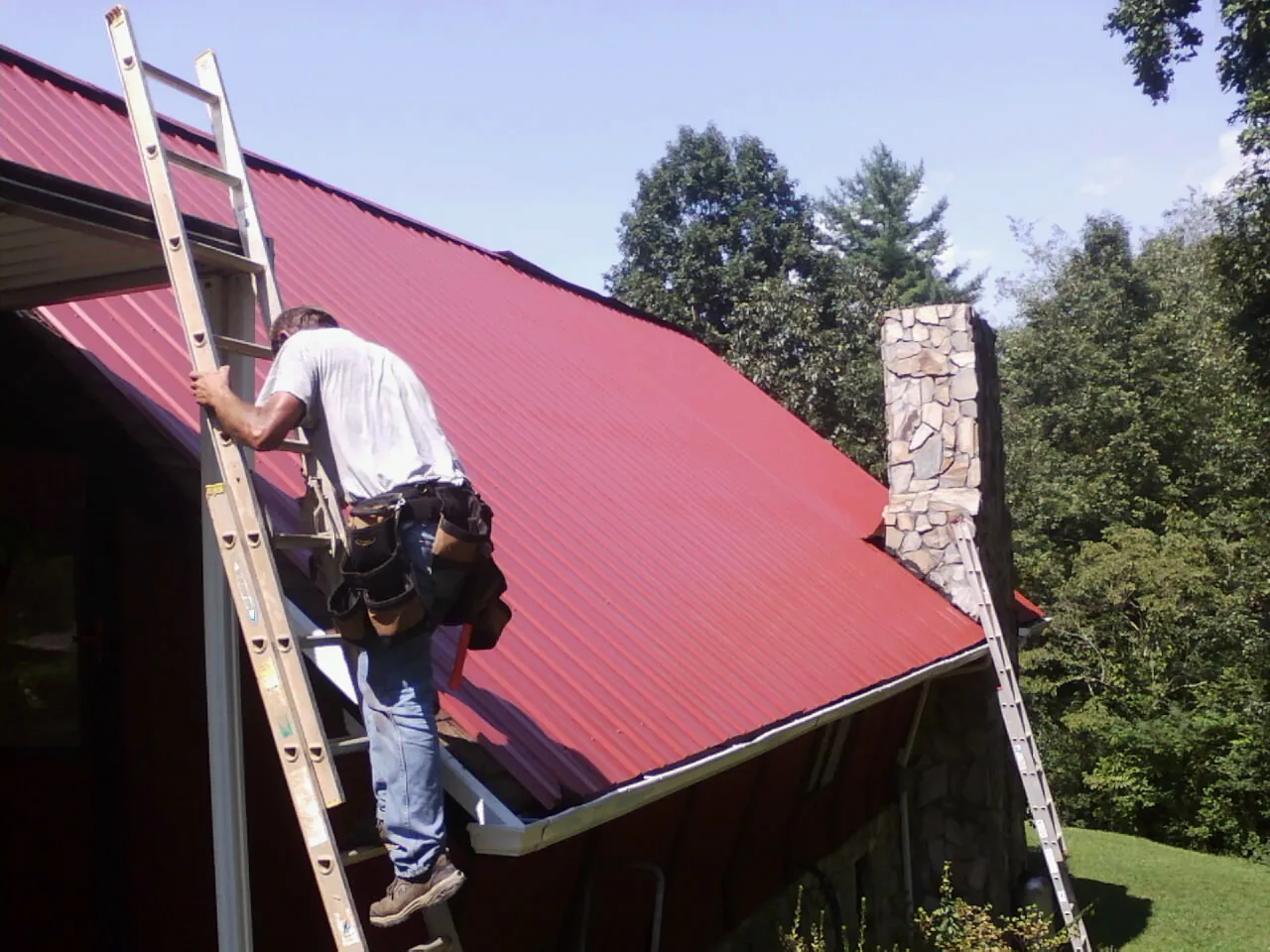 Roofer on a ladder beside a red metal roof and stone chimney — Soffit & Fascia Repair by Roofing Brothers in Visalia